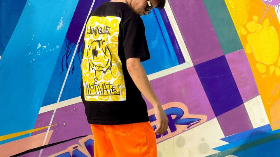 Person wearing a black t-shirt with yellow graphic and orange shorts standing in front of a colorful mural.